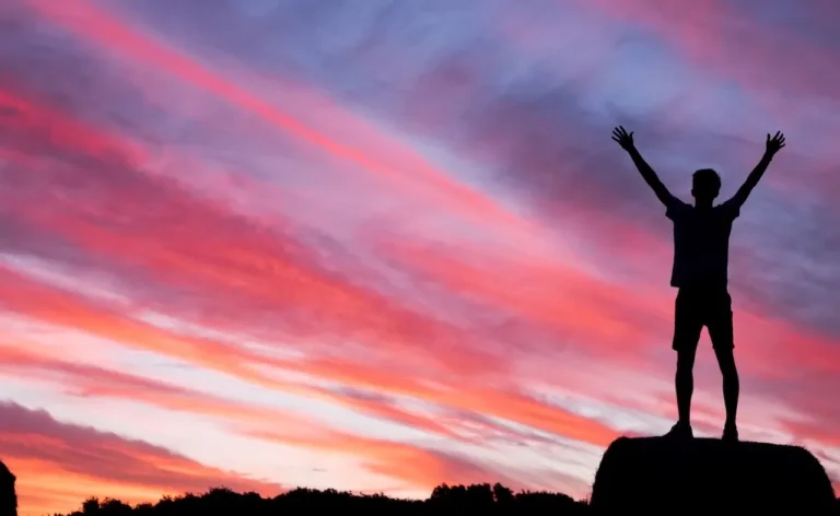 Image of someone standing on a mountain top, raising their arms, against a sunset sky
